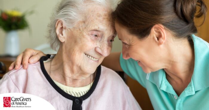 A caregiver lovingly embraces a senior woman while they both smile, representing the kind of warm disposition that can help when dealing with dementia-related repetition.