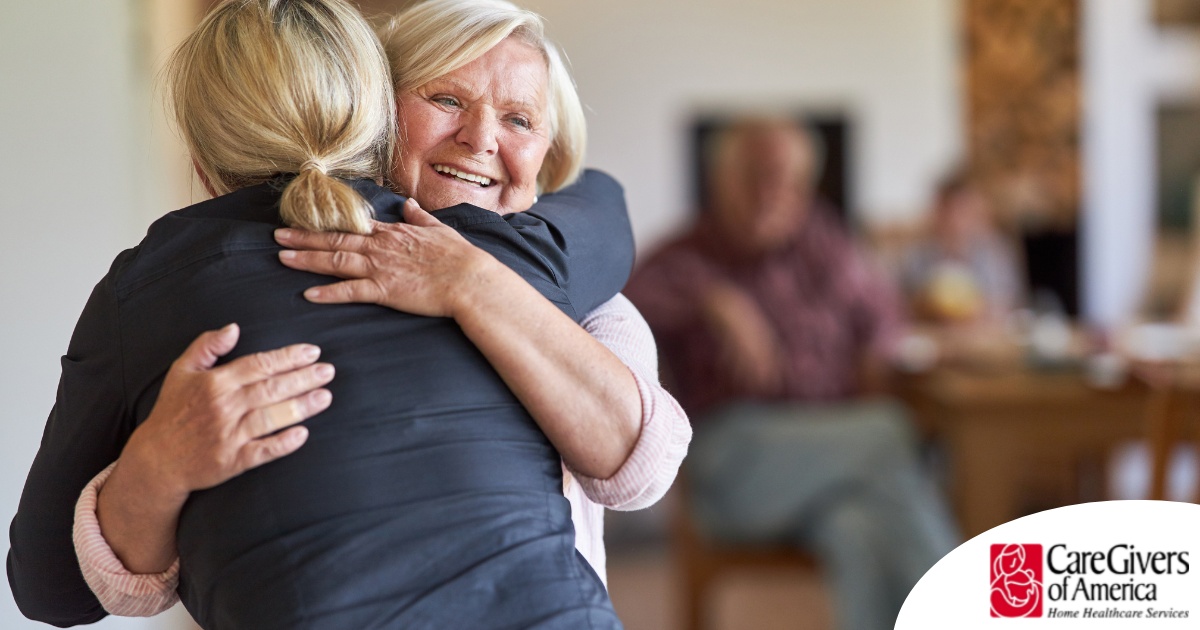 An older woman smiles as a younger woman visits her and hugs her, showing the effect that acts of kindness can have on senior loved ones.