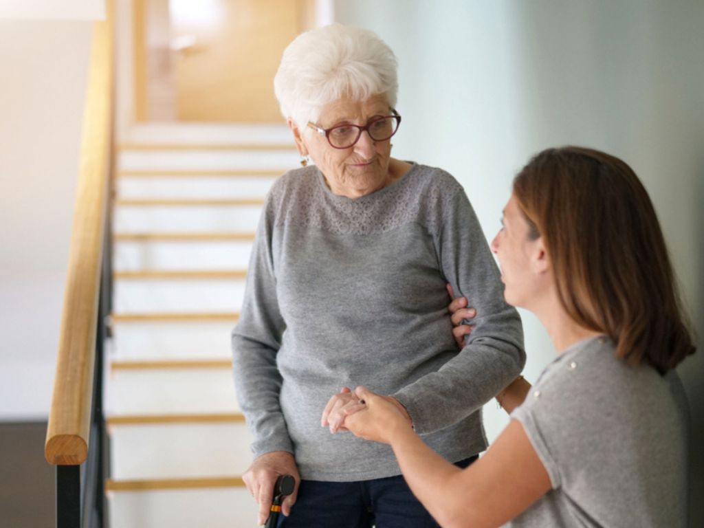 A caregiver helps an elderly woman get down the stairs. In-Facility Care in Aventura