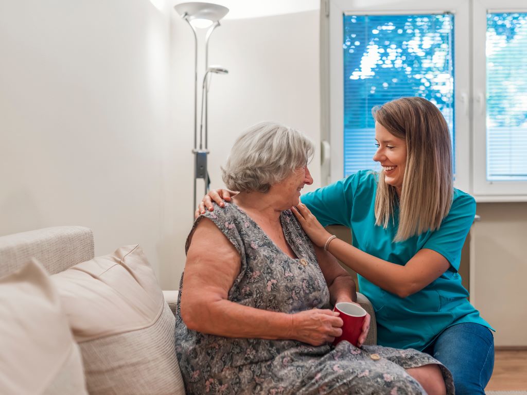 A cheerful care giver sits on a couch next to an elderly patient laughing
