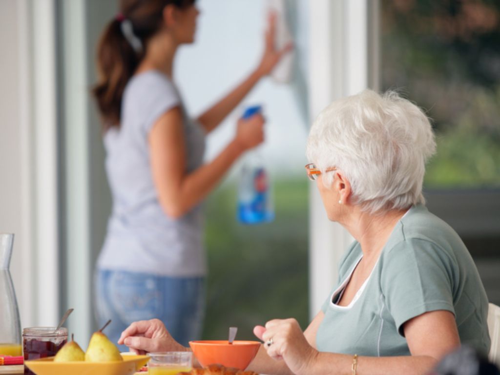 An elderly woman side at a table outside, in the background a care giver cleans a window. Light Housekeeping in Cooper City