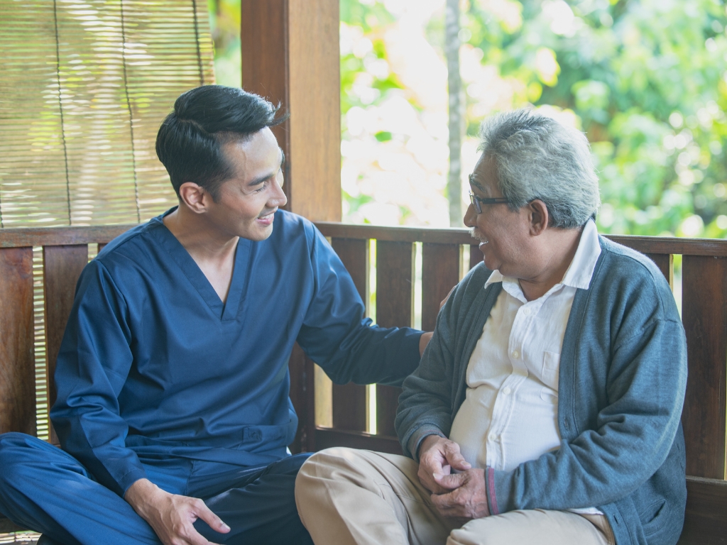 A cheerful care giver sits next to an elderly gentleman on a bench as they talk to each other. Senior Home Care in Broken Sound