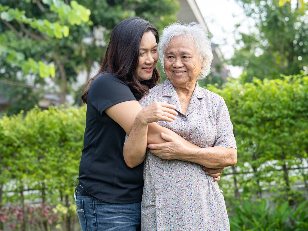 Young Asian woman embracing an elderly Asian woman outdoors in a green, sunny setting, both smiling brightly and radiating happiness and warmth.