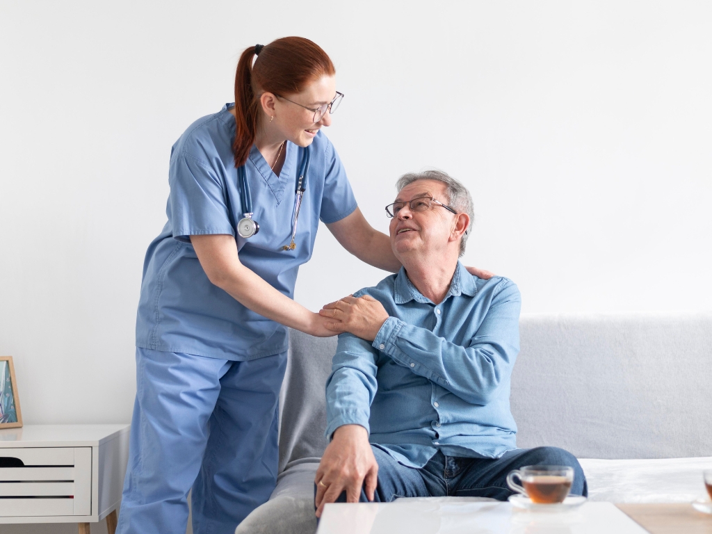 A young female caregiver in blue scrubs stands behind a senior man seated on a sofa, gently resting her hands on his shoulders as he looks up at her with a smile.