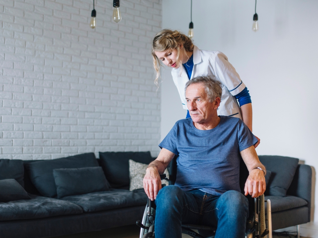 Young caregiver standing behind an elderly man as they both look in the same direction in a cozy living room, sharing a quiet and thoughtful moment.