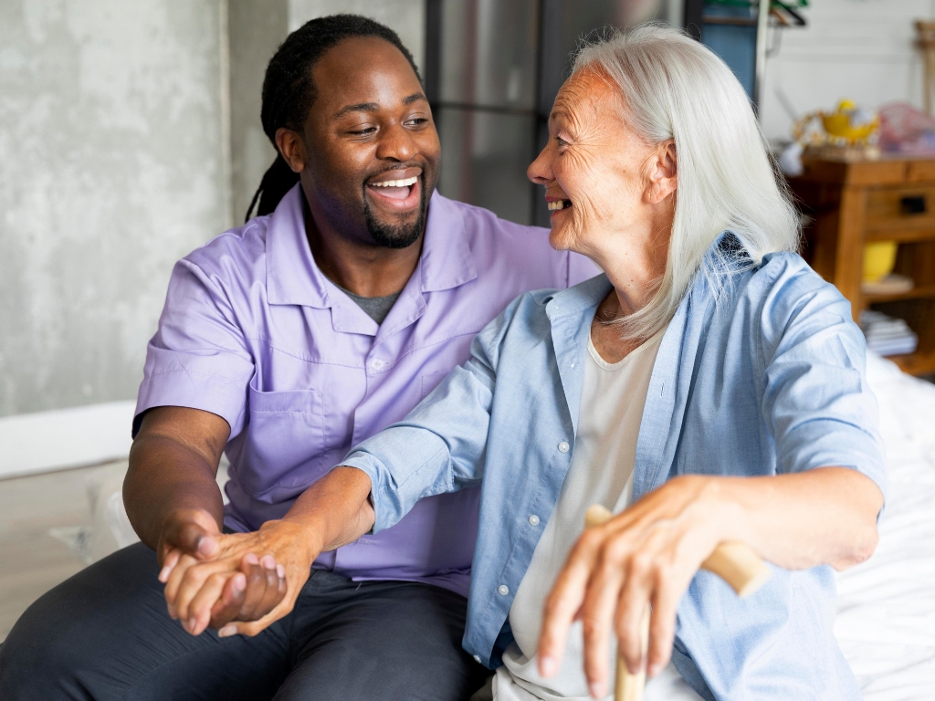 A black male caregiver with long beaded hair tied in a ponytail gently places his hand over the hand of a senior woman, both of them laughing and sharing a joyful moment while looking at each other in Vero Beach.