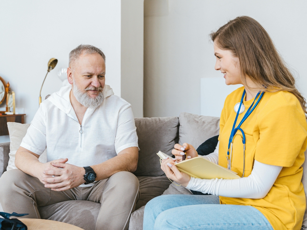 A female caregiver talks with an older man on a couch in a warm home setting, reflecting friendly companion care in Boynton Beach.