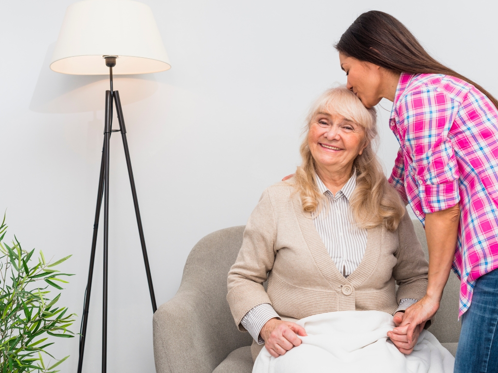 Alt image description: A smiling elderly woman sits comfortably in a chair while a younger woman gently kisses her on the forehead and holds her hand. The warm and caring interaction reflects the compassionate support provided through senior home care in Indian River County.