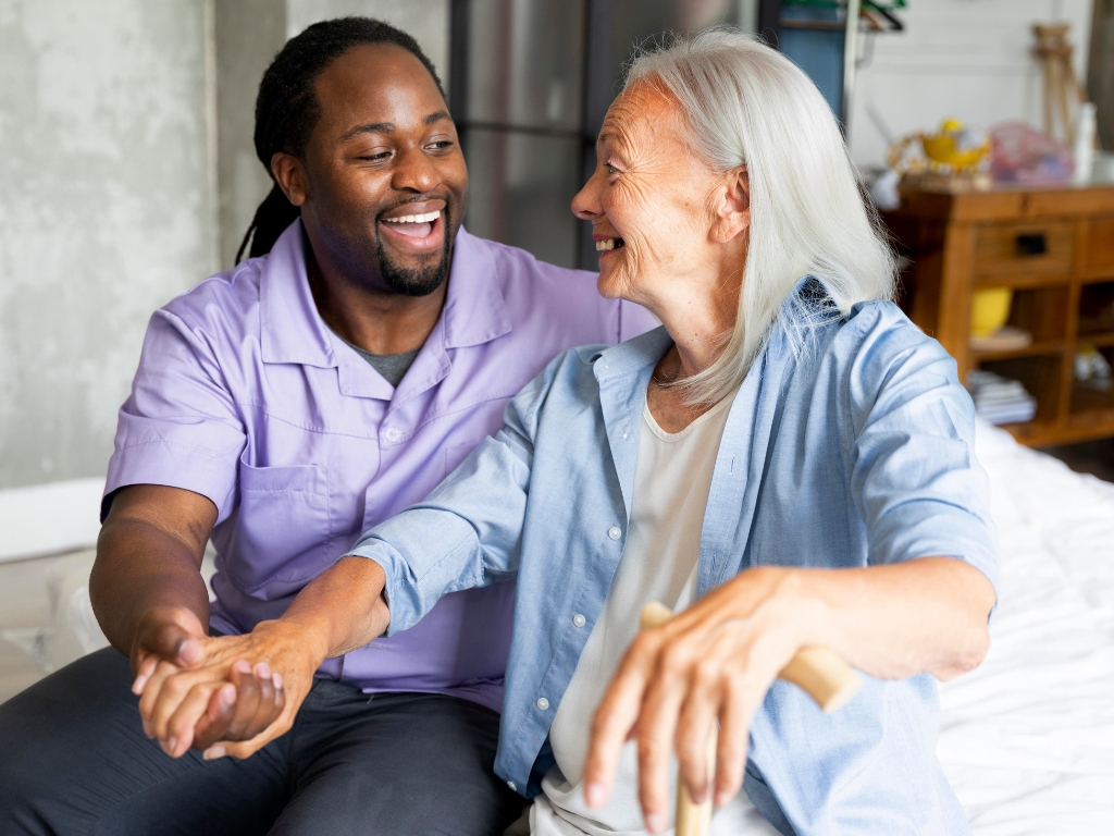 A Black male caregiver sits closely with a senior woman, both smiling and looking at each other with warmth and trust. Their connection reflects the compassionate and personalized support offered through home care in Palm City.