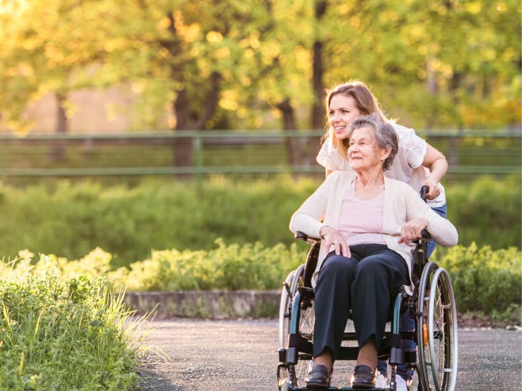 Senior woman in a wheelchair with a female caregiver standing behind her, both looking thoughtfully into the distance outdoors, representing attentive Live-In and 24-Hour Care