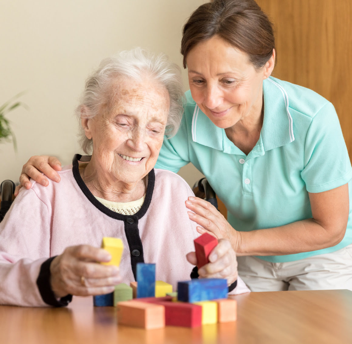 building-puzzles (1) Female caregiver assisting a senior woman with building blocks at a table, engaging her in a cognitive activity to support dementia care with patience and encouragement