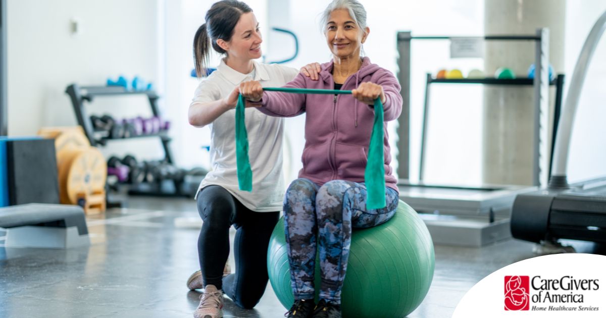 A care provider helps an older woman exercise with a resistance band and an exercise ball, representing how exercise can help with senior fall prevention.
