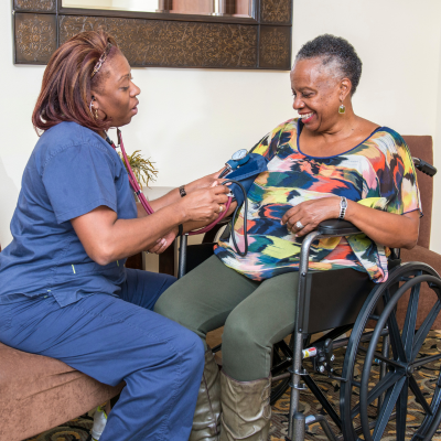 A caregiver in blue scrubs checks the blood pressure of a smiling woman sitting in a wheelchair during an in-home care visit.