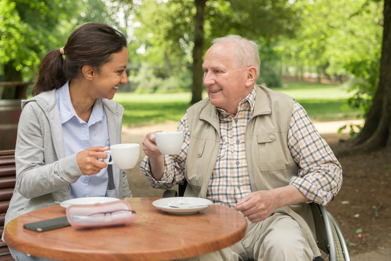 A smiling caregiver and a senior man in a wheelchair sharing coffee at an outdoor park, representing dependable live-in and 24-hour care in Margate, FL.