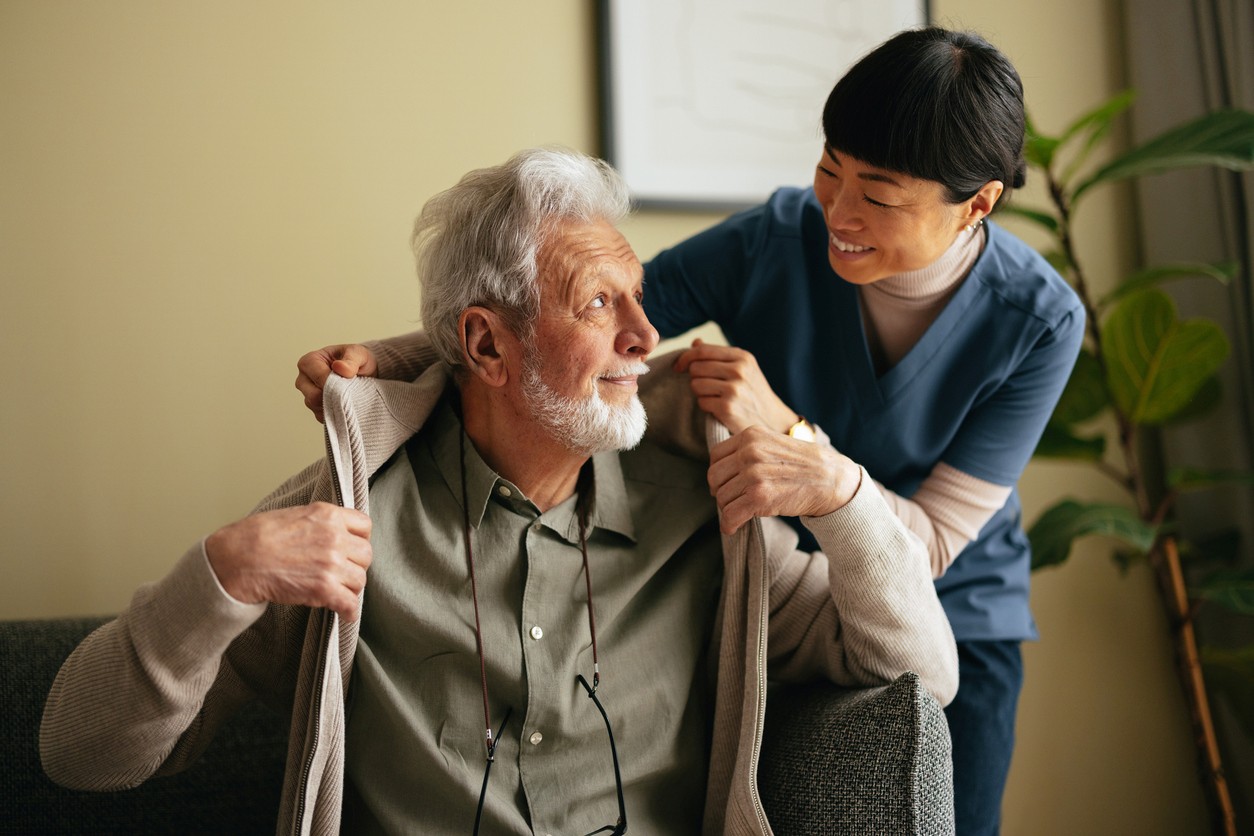 A kind caregiver in blue scrubs assisting a smiling elderly man with his sweater, showcasing reliable respite care in Margate, FL.